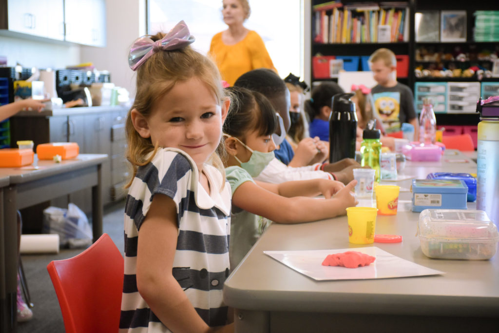 a young girl sitting at her desk in a classroom setting; in the background, students mold Play-Dough - link opens photo in full resolution