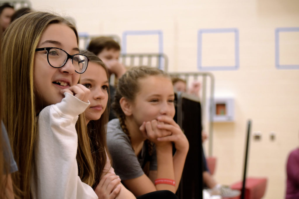 three students watching a school assembly from the bleachers in the gym - link opens photo in full resolution